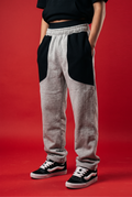 A person wearing heather gray sweatpants with large black color-block panels on the upper thighs, paired with black and white Vans sneakers, set against a solid red studio background.