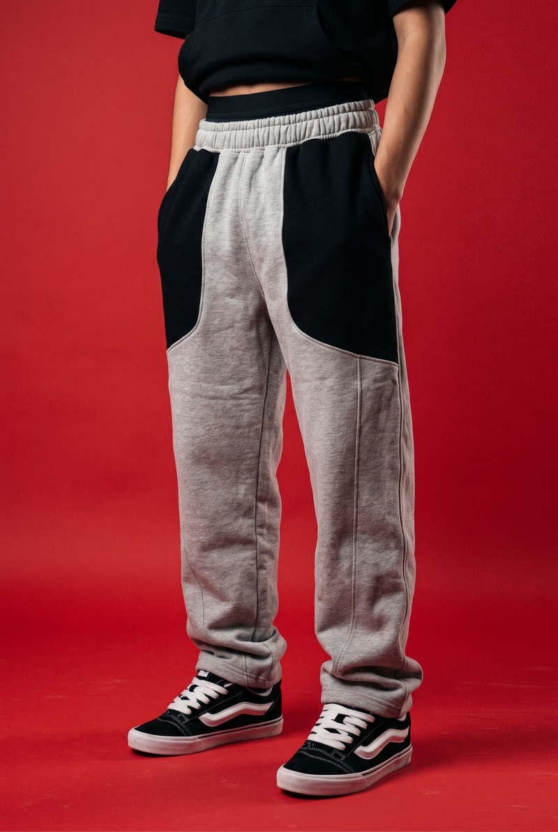 A person wearing heather gray sweatpants with large black color-block panels on the upper thighs, paired with black and white Vans sneakers, set against a solid red studio background.