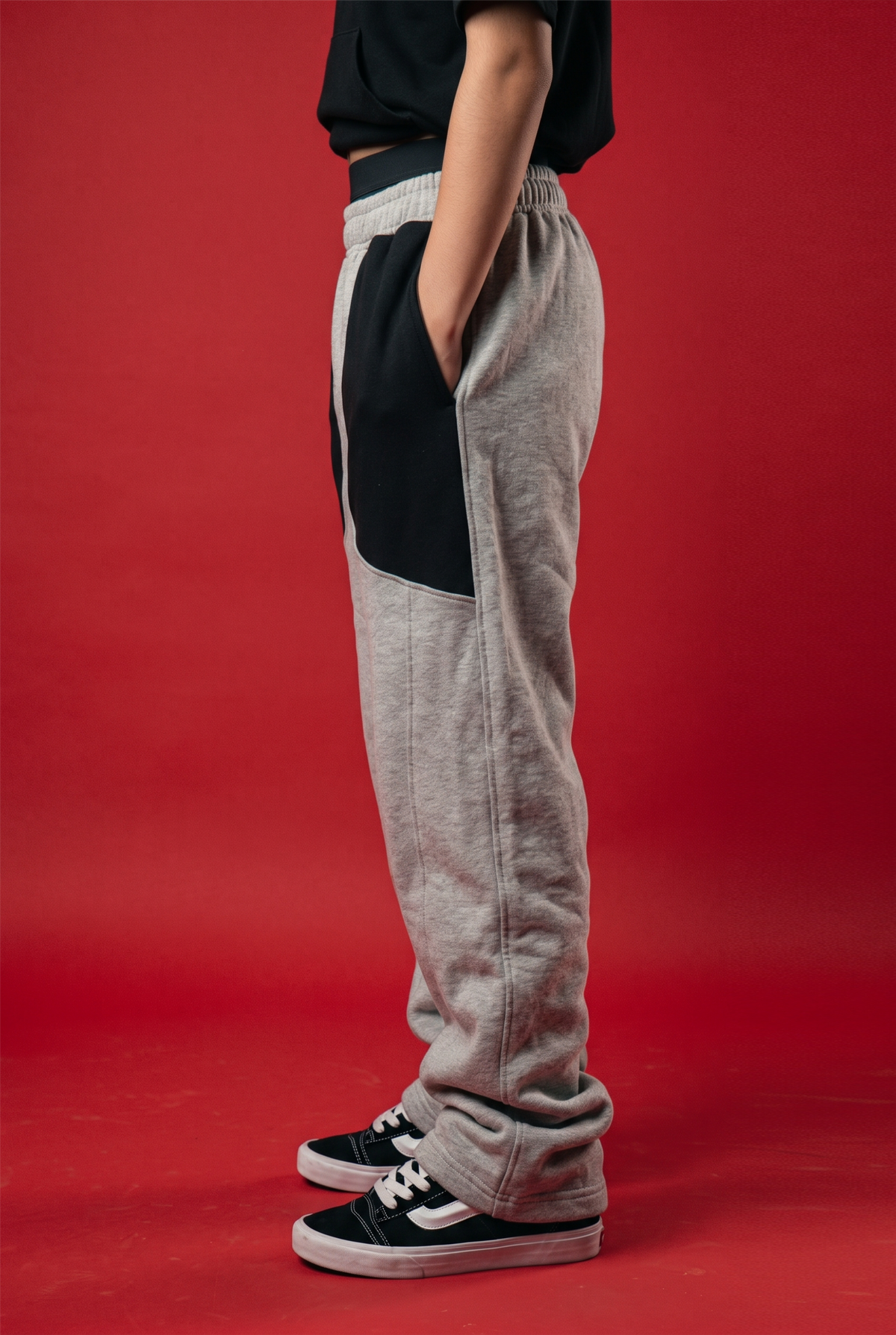A person shown from the waist down wearing heather gray sweatpants with black side panels and a black waistband, paired with a black top and black low-top sneakers, against a solid red studio background.
